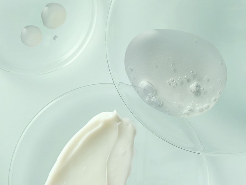 Dermocosmetic skincare textures showing a smear of white cream, a blob of clear gel with bubbles, and serum drops on glass petri dishes in a lab setting.