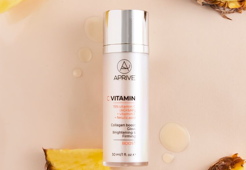 APRIVE C Vitamin Boost serum bottle containing 15% vitamin C, vitamin E, and ferulic acid, surrounded by serum drops and pineapple slices.
