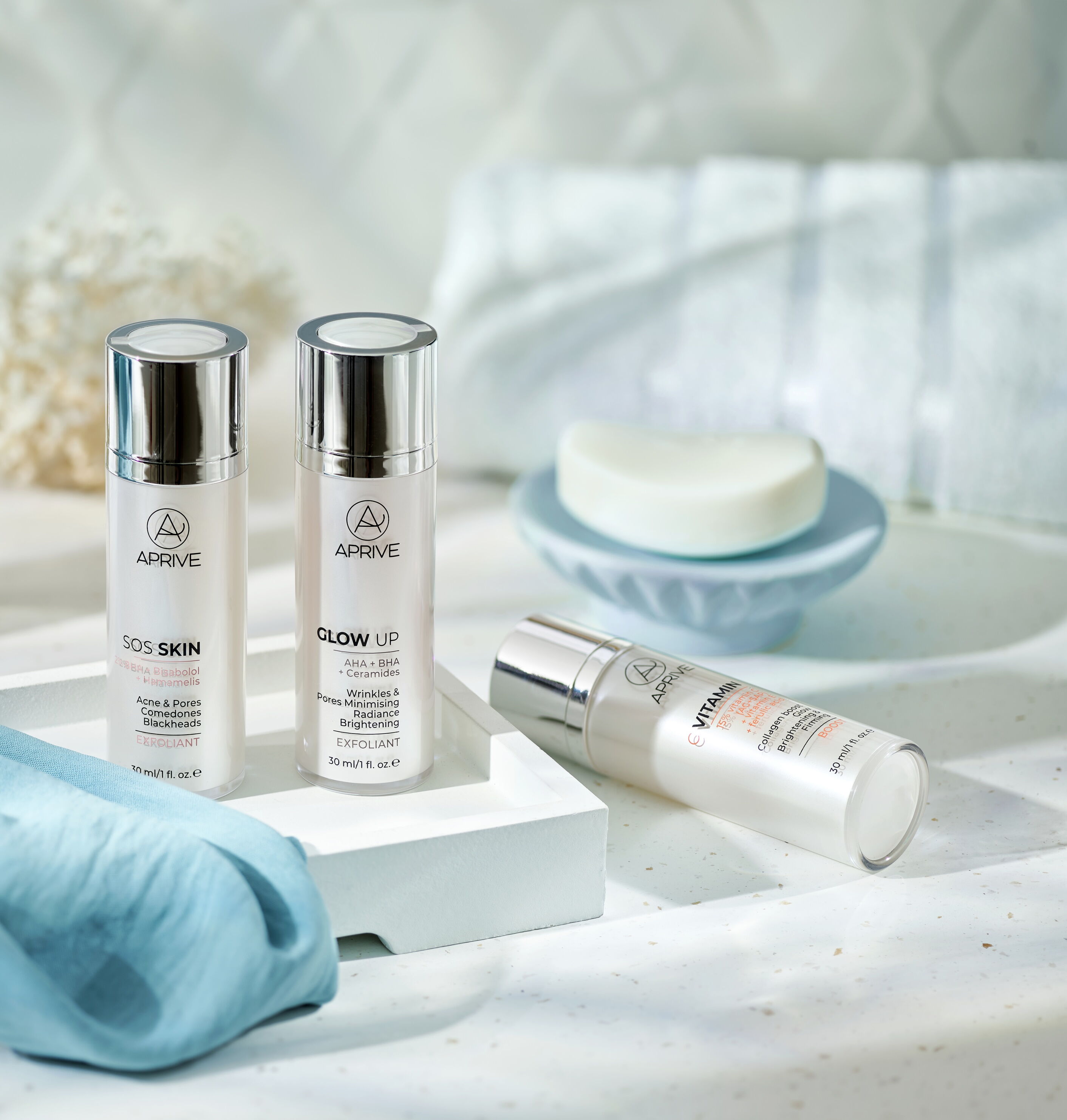 Tri bočice Aprive proizvoda za njegu kože na bijelom pladnju u kupaonici: SOS Skin eksfolijant za akne, Glow Up AHA+BHA eksfolijant za bore i sjaj, te Vitamin C Booster serum.