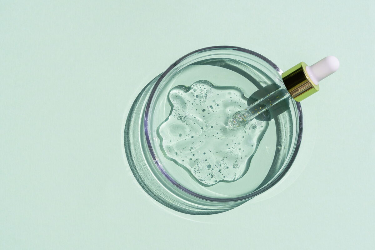 Top-down view of a glass petri dish containing a transparent, bubbly cosmetic gel serum and a gold-top dropper on a light green background.