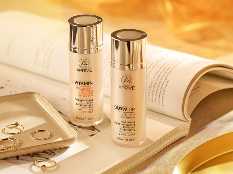 APRIVE Vitamin C brightening serum and Glow Up AHA BHA exfoliant bottles displayed on an open book with gold jewelry.