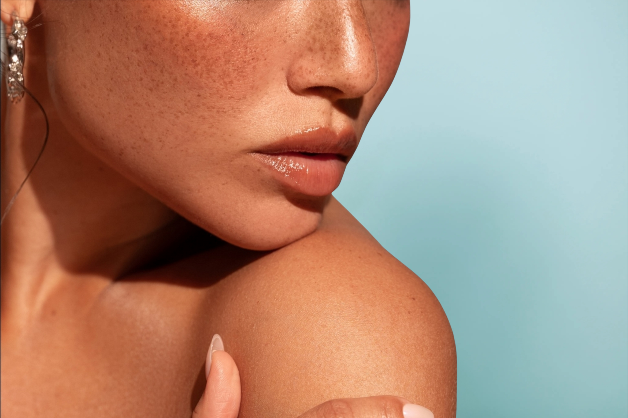 Close-up view of a woman with freckles and glowing skin wearing Aprive Cosmetics lip gloss against a blue background.