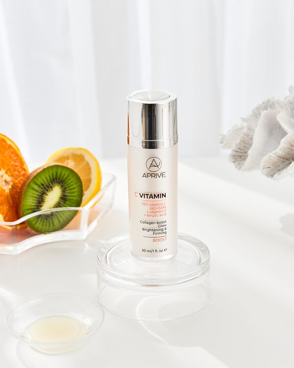 Bottle of Aprive 15% Vitamin C Boost serum on a glass stand with fresh orange and kiwi slices in the background.