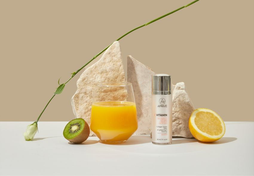 Bočica Aprive Vitamin C Seruma pored čaše soka od naranče, polovice kivija i polovice limuna, okružena prirodnim kamenjem i zelenom stabljikom.