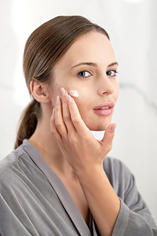 A young woman with brown hair tied back is applying a white moisturizing cream to her cheek with her fingertips in a bright bathroom.