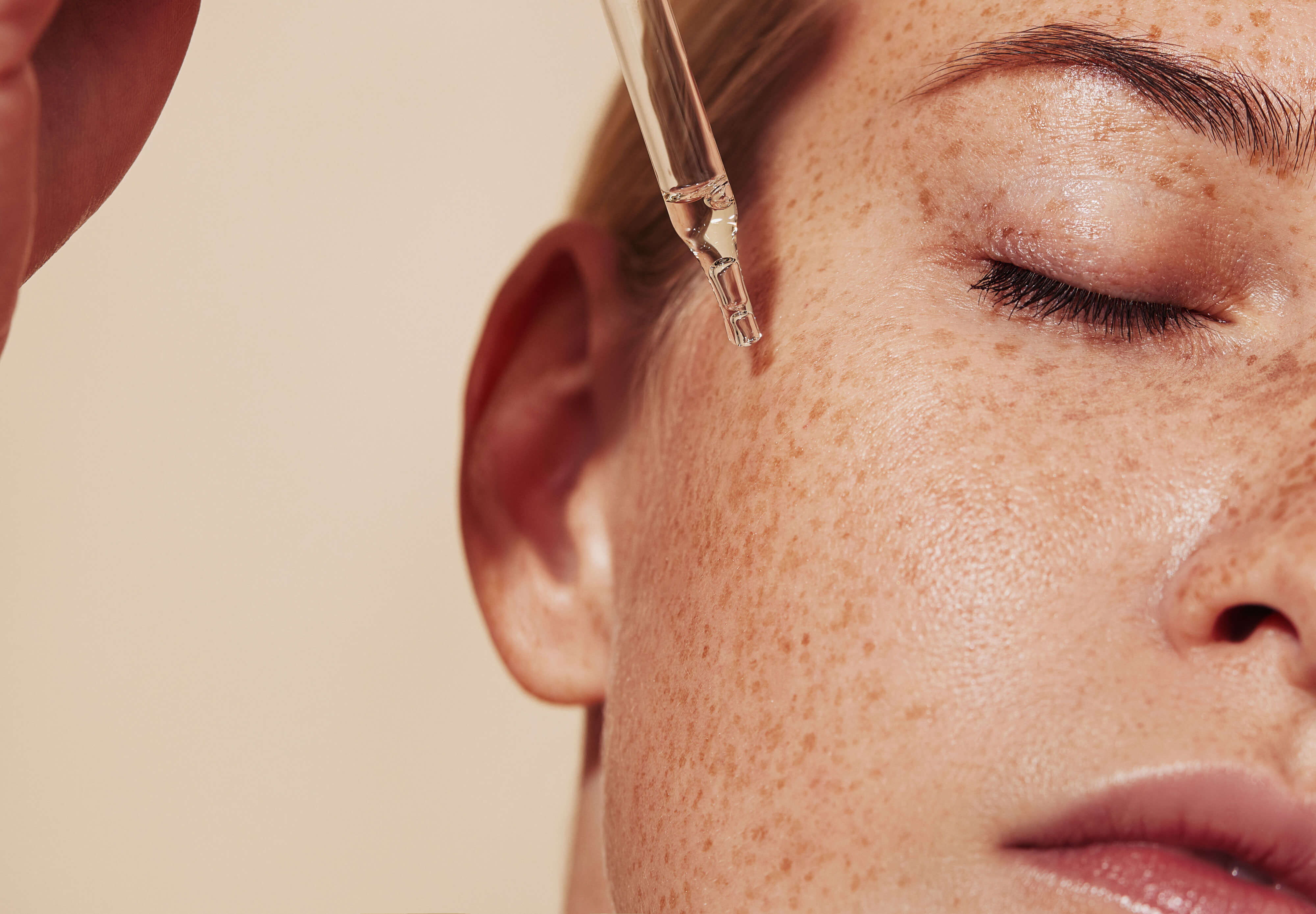 Close-up view of a dropper dispensing clear facial serum onto a woman's cheek covered in freckles with her eyes closed.