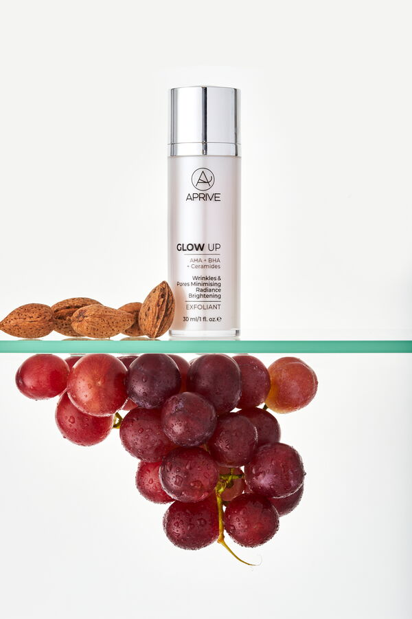 Aprive Glow Up Exfoliant bottle on a glass shelf alongside almonds and red grapes, indicating natural ingredients like AHA and BHA.