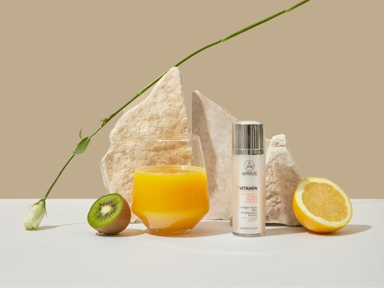 Bottle of Aprive Vitamin C Serum next to a glass of orange juice, a halved kiwi, and a halved lemon, surrounded by natural stones and a green stem.