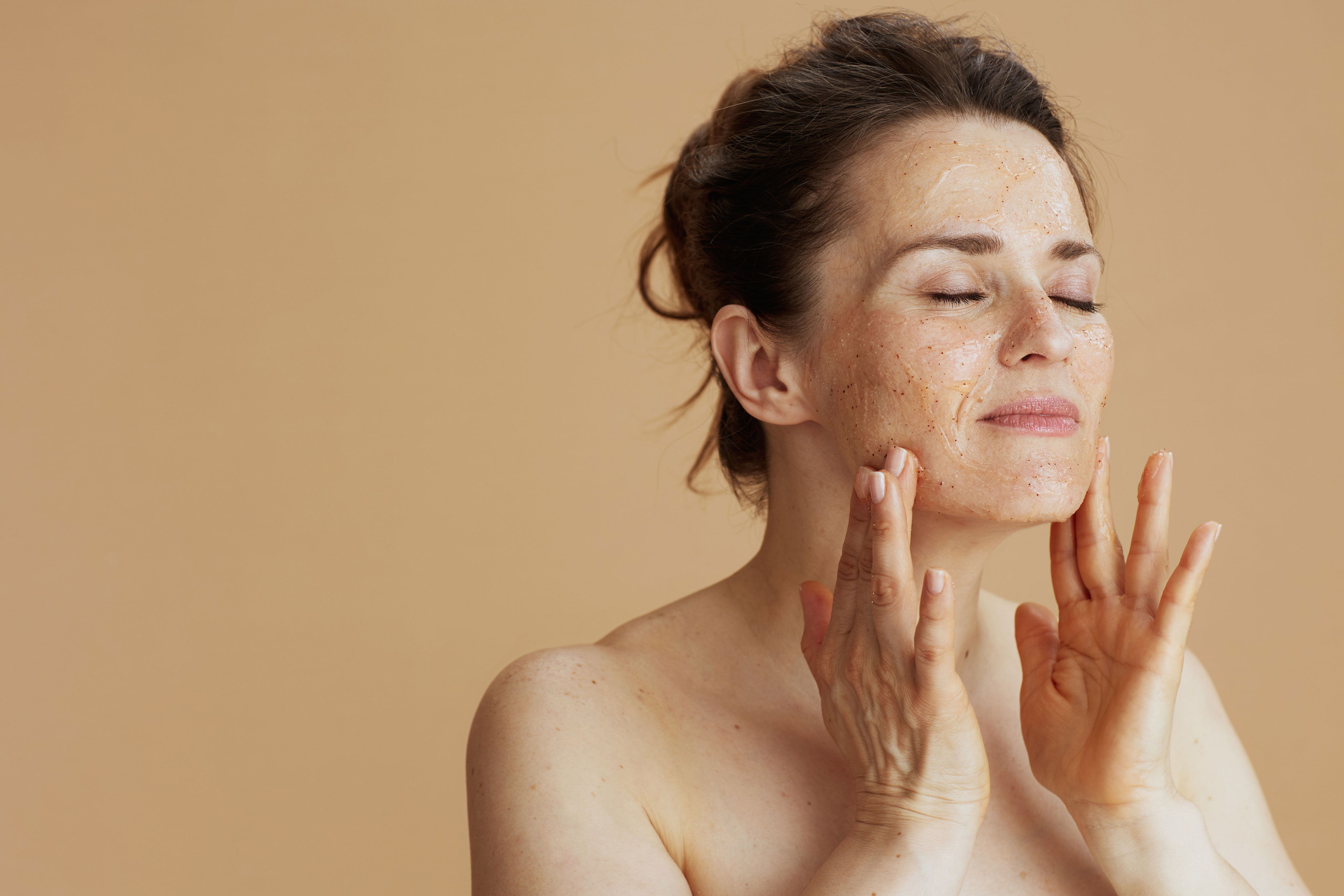 A middle-aged woman with her eyes closed, gently applying a natural exfoliating facial scrub to her cheeks against a beige background.