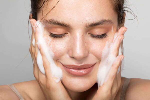 Close-up of a smiling woman with freckles massaging white foamy cleansing gel onto her cheeks with both hands.