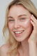 A close-up portrait of a smiling blonde woman applying white moisturizing cream to her cheek.