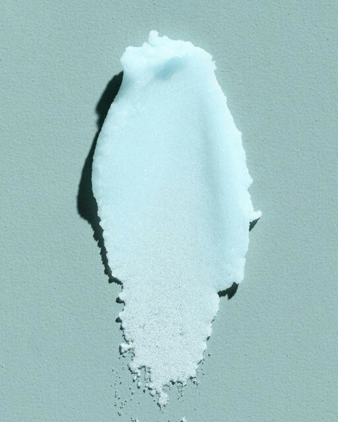 A close-up photograph of a vertical smear of granular blue shea butter body scrub on a pale blue background, showing its texture.