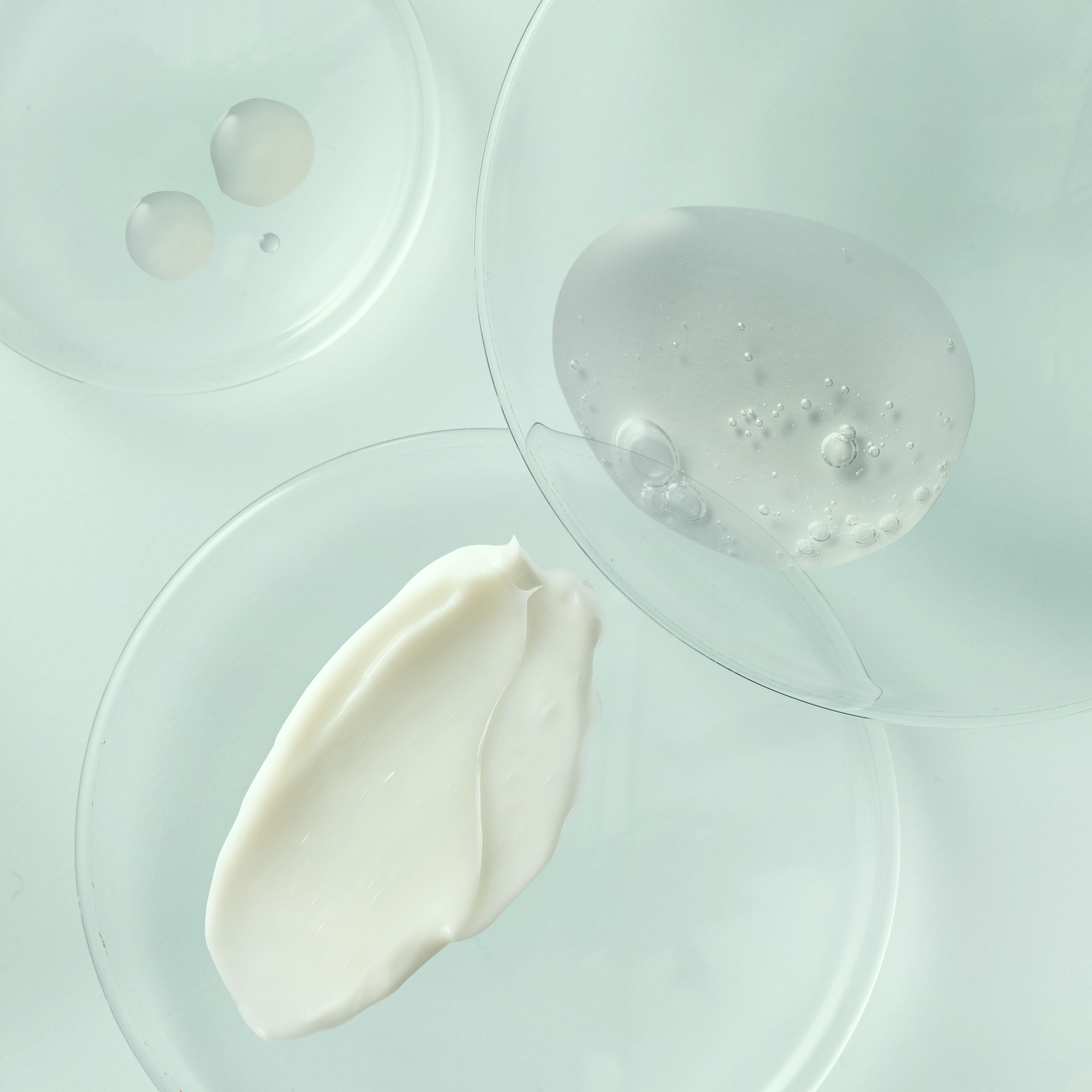 Overhead view of skincare textures in glass petri dishes, showing white cream, clear gel with bubbles, and translucent serum drops on a light blue background.