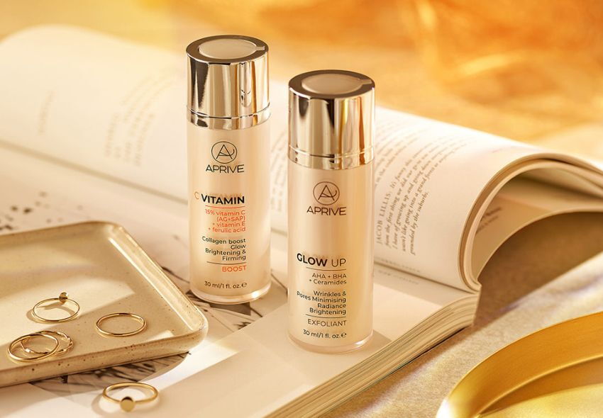 APRIVE Vitamin C brightening serum and Glow Up AHA BHA exfoliant bottles displayed on an open book with gold jewelry.