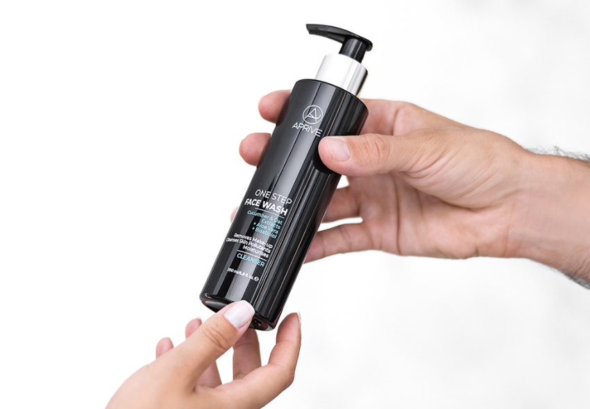 A person's hands holding a black pump bottle of Aprive One Step Face Wash cleanser.