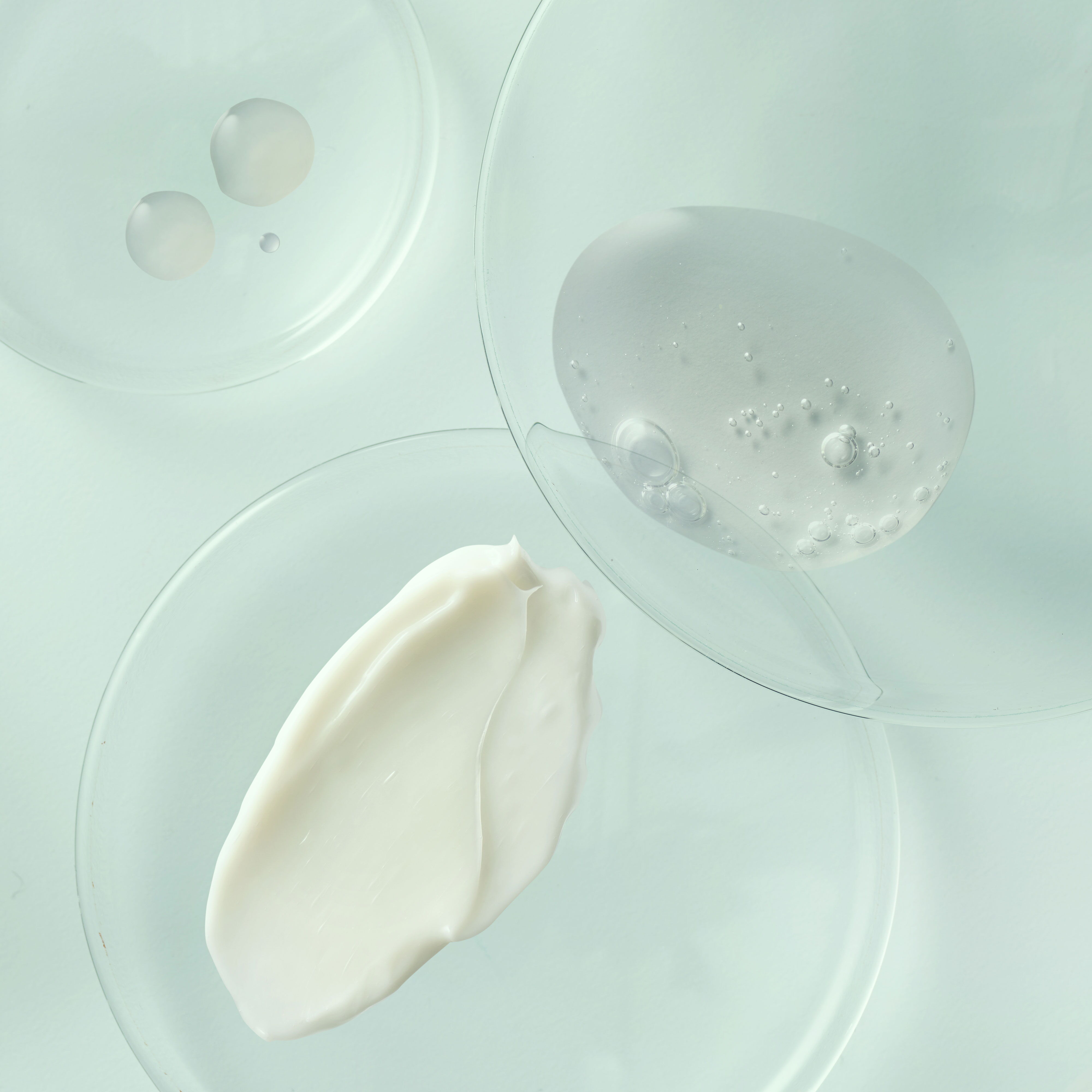 Top-down view of cosmetic creams, gels, and serums in glass petri dishes for skincare research on a light blue background.