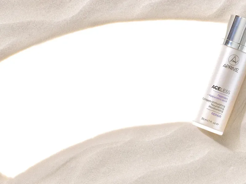 A bottle of Aprive Ageless Peptides + Hyaluronic Acid Repair cream resting on sand next to a smear of the product texture.