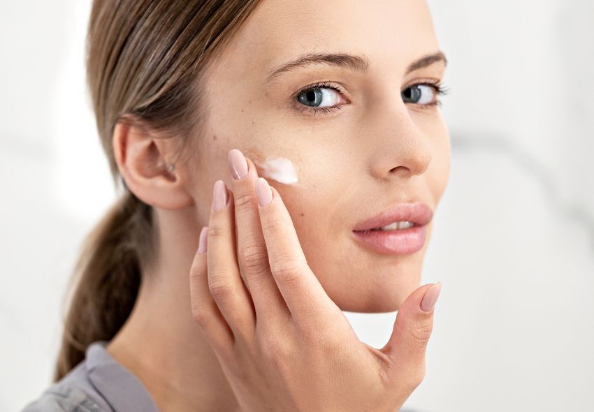 Žena čiste kože nježno nanosi bijelu retinol kremu na obraz kao dio njege lica.