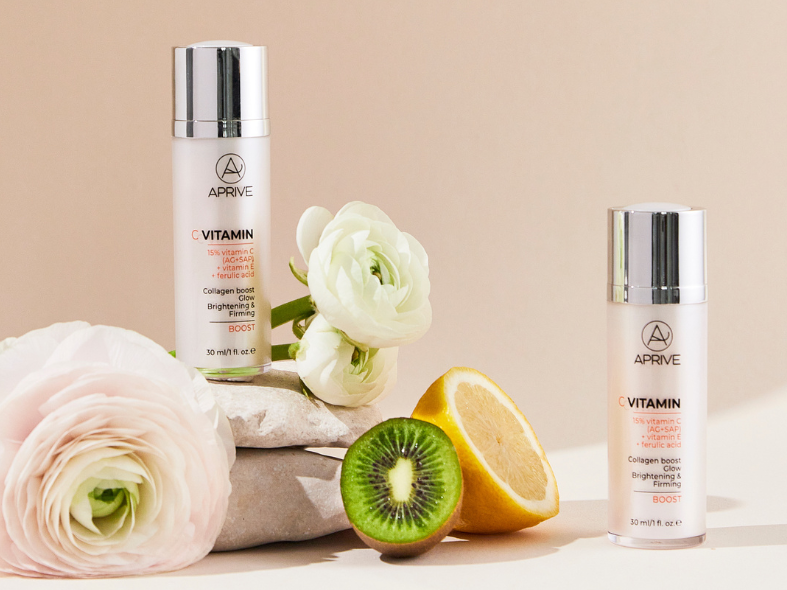 Two bottles of APRIVE 15% Vitamin C serum with Vitamin E and Ferulic Acid, displayed with fresh kiwi, lemon halves, and white ranunculus flowers on stones.