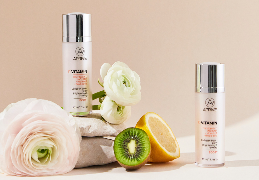 Dvije bočice APRIVE 15% seruma s vitaminom C, vitaminom E i ferulinskom kiselinom, prikazane sa svježim kivijem, polovicama limuna i bijelim cvjetovima žabnjaka na kamenju.